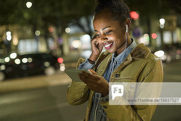 Portrait of smiling young woman using earphones and smartphone in the city by night  Lisbon  Portugal