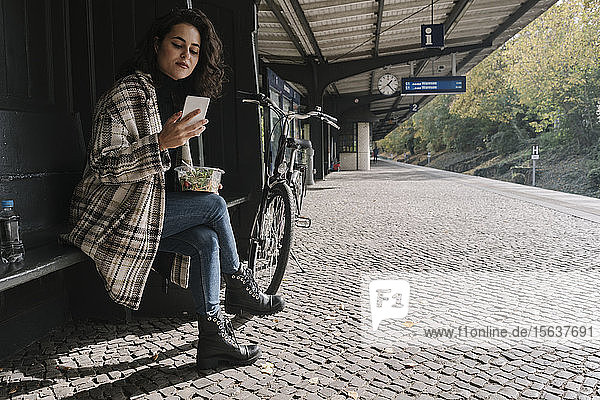 Frau mit Fahrrad beim Mittagessen und mit Smartphone auf dem Bahnsteig  Berlin  Deutschland