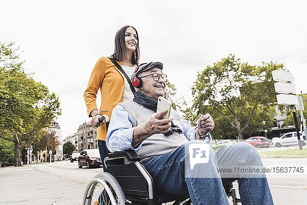 Junge Frau schiebt älteren Mann mit Kopfhörern und Smartphone im Rollstuhl