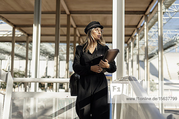 Junge blonde Geschäftsfrau mit schwarzer Matrosenmütze und Laptoptasche in der Hand