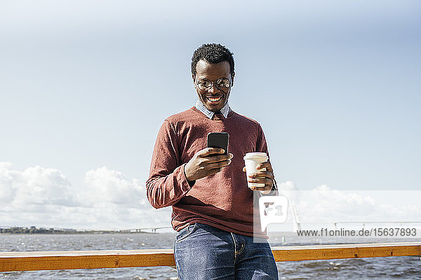 Junger Mann steht am Meer  hält Kaffee in der Hand und benutzt ein Smartphone