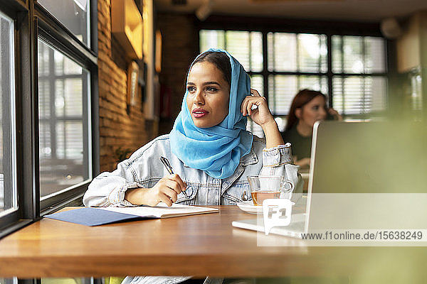 Geschäftsfrau  die in einem Café einen türkisfarbenen Hidschab trägt und in ein Notizbuch schreibt