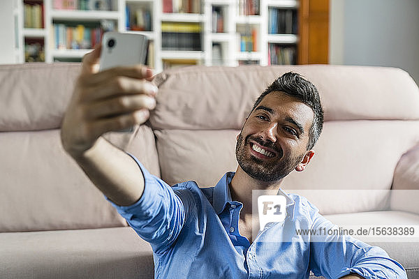 Portrait of smiling young man sittingin front of couch at home taking selfie with cell phone