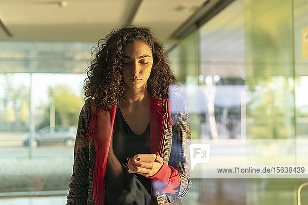 Porträt einer jungen Frau hinter Glasscheibe mit Blick auf Handy