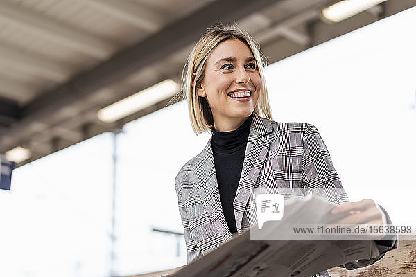 Lächelnde junge Geschäftsfrau mit Zeitung am Bahnhof