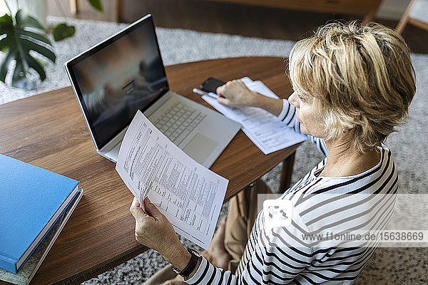 Reife Frau mit Dokumenten,  die zu Hause einen Laptop benutzen