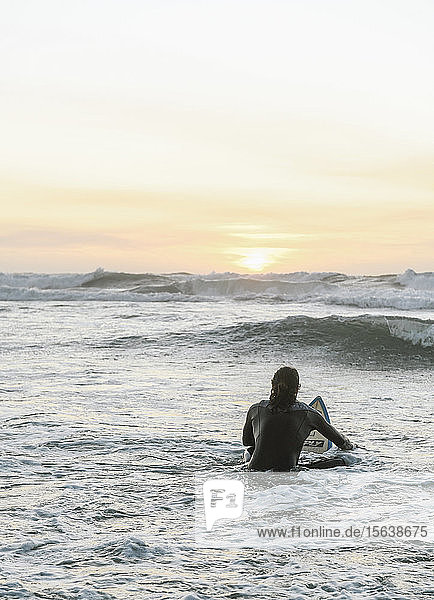 Rückansicht eines Surfers mit Surfbrett  der während des Sonnenuntergangs auf einem Surfbrett im Meer sitzt