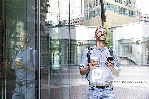 Lächelnder Geschäftsmann mit Tasse Kaffee und Smartphone in der Hand in der Stadt  Berlin  Deutschland