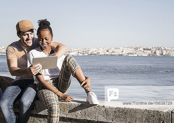 Happy young couple sitting on a wall at the waterfront using a tablet  Lisbon  Portugal