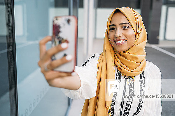 Junge muslimische Frau  die einen gelben Hidschab trägt  ein Smartphone benutzt und ein Selfie