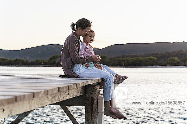 Glückliche Mutter mit Tochter sitzt bei Sonnenuntergang auf einem Steg