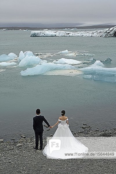 Braut in einem klassischen weißen Hochzeitskleid und Bräutigam in einem dunklen Anzug mit Blick auf schwimmende Eisblöcke und den Gletscher am Ufer der Lagune Jökulsárlón  Jökulsárlón  Vatnajökull-Nationalpark  Südisland  Island  Europa