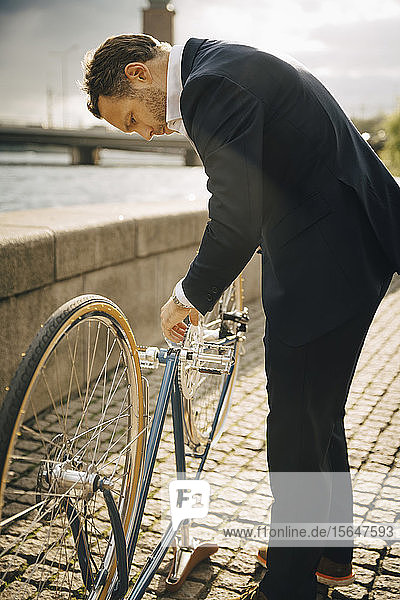 Side view of businessman repairing bicycle on footpath in city during sunny day