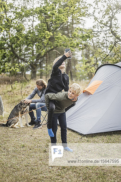 Junge nimmt Schwester mit Smartphone huckepack  während Mann beim Zeltcamping mit Hund spielt