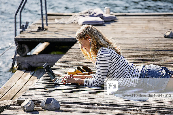 Mid adult female typing on laptop while lying over wooden pier during summer vacation