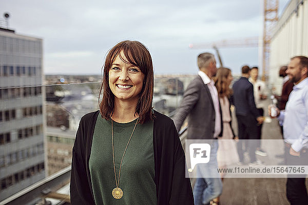 Porträt einer lächelnden Geschäftsfrau mit Kollegen im Hintergrund  die während der Party auf der Terrasse stehen