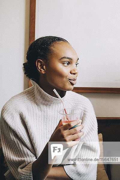 Smiling teenage girl looking away while having smoothie at home