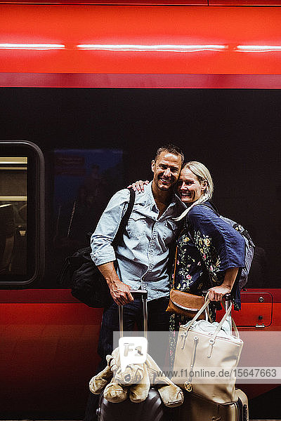 Porträt eines glücklichen Paares mit Gepäck  das am Bahnhof gegen den Zug steht