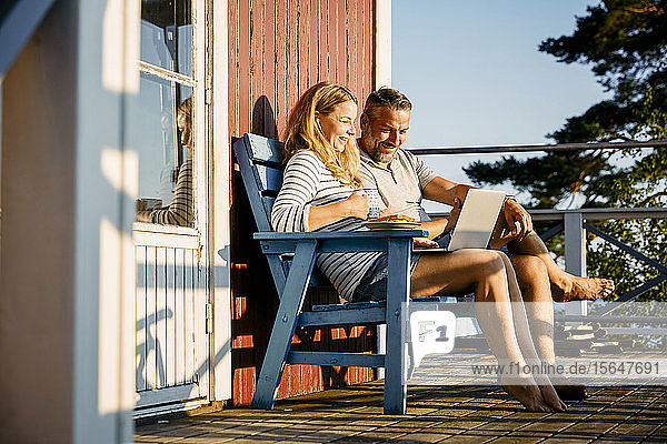 Lächelndes Paar mit Laptop beim Frühstück auf der Veranda im Blockhaus