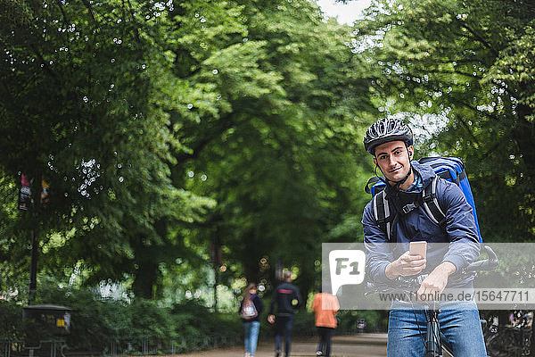 Portrait of confident delivery man with bicycle against trees in city