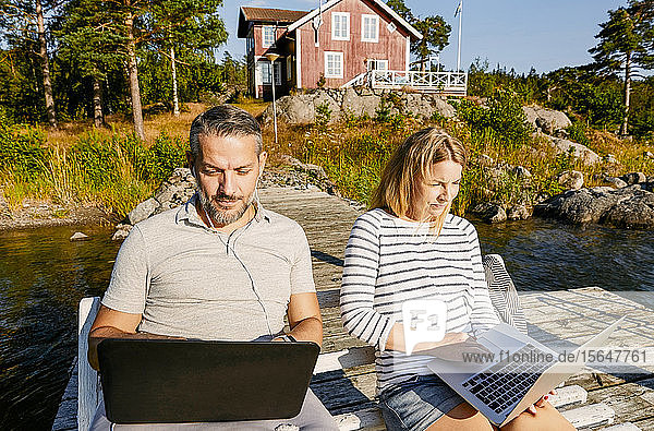 Couple using laptop while sitting on bench together at pier during summer