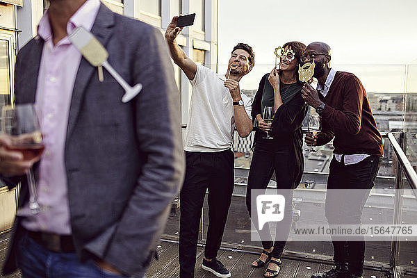 Office friends with props taking selfie while enjoying drinks at party on terrace