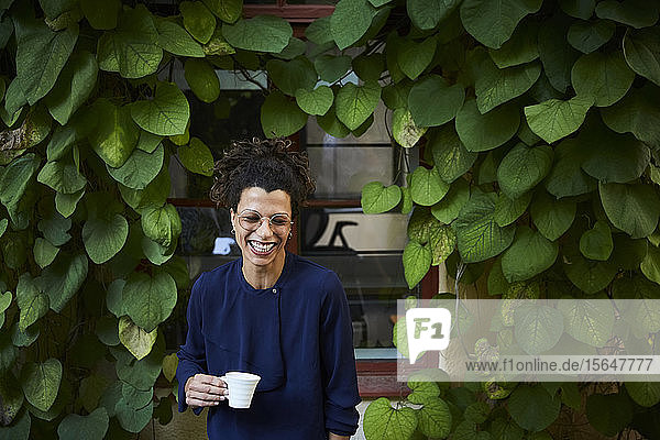 Glückliche junge Architektin hält Kaffeetasse gegen Kletterpflanzen im Garten