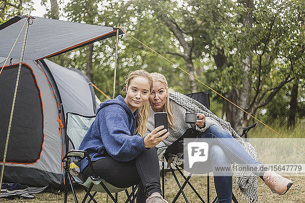 Mother and daughter taking selfie on smart phone while having coffee in campsite