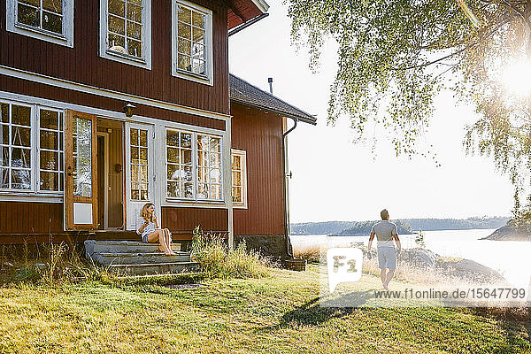 Woman sitting on steps at entrance of log cabin while man walking towards lake on sunny day