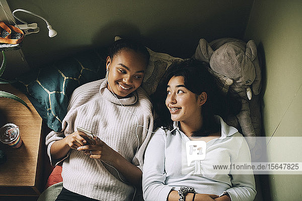 Directly above shot of smiling teenage girls listening music on mobile phone while lying on bed at home