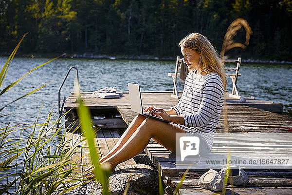 Frau mit Kopfhörern  die einen Laptop benutzt  während sie auf einem Steg am See sitzt