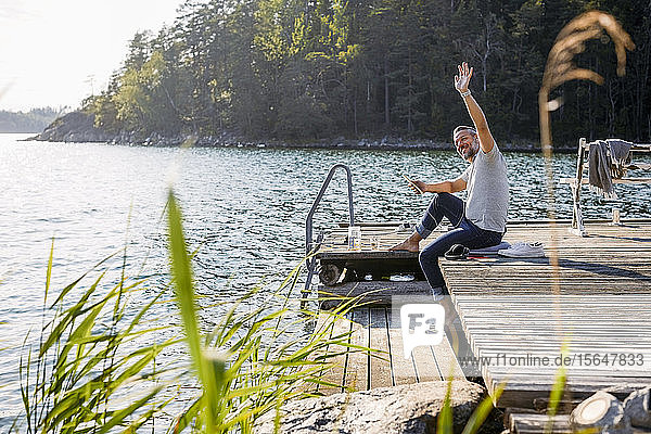 Mature man looking away and waving while holding digital tablet on jetty over lake