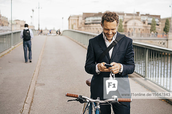 Smiling businessman using mobile phone while standing with bicycle on bridge in city