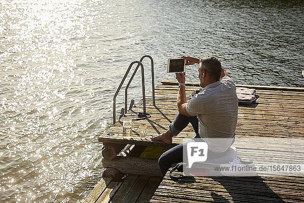 High angle view of mature male photographing on digital tablet while sitting on pier over lake