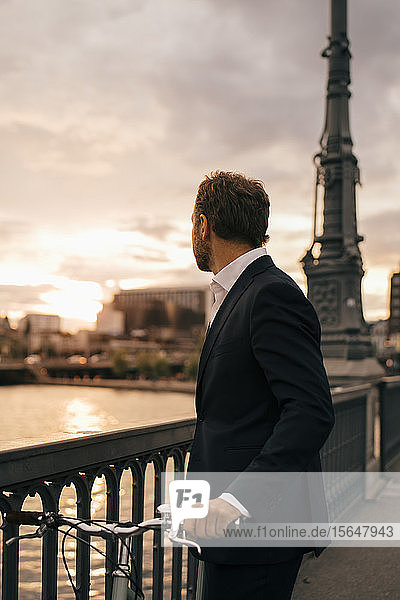 Side view of businessman with bicycle standing on bridge in city during sunset