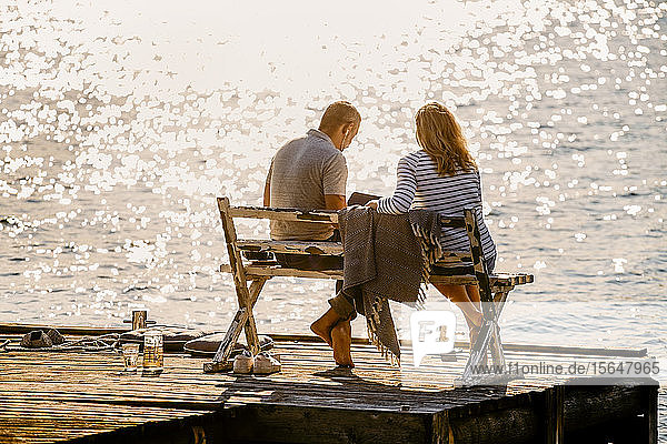 Paar in voller Länge mit Laptop  während es in den Sommerferien auf einer Bank über dem Holzsteg am See sitzt