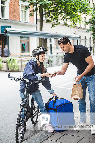 Zustellerin nimmt Zeichen eines männlichen Kunden entgegen  während sie ein Paket auf der Straße in der Stadt ausliefert