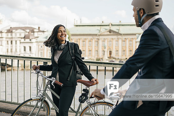 Smiling businesswoman with bicycle looking at colleague biking on bridge