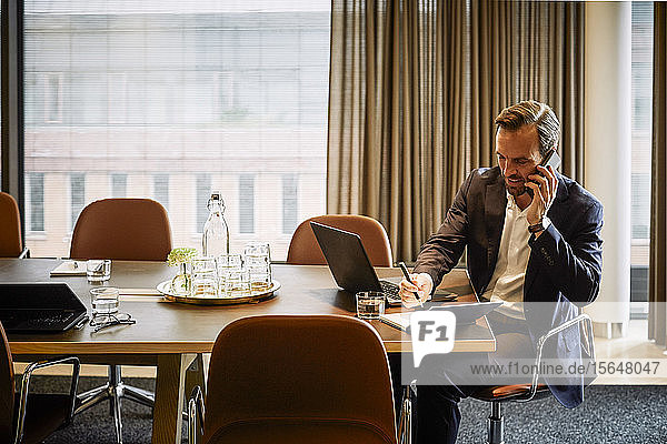 Confident male lawyer talking on mobile phone while planning in diary at board room