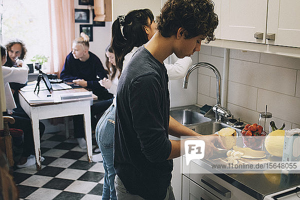 Teenage boy with female cutting fruits at kitchen counter while friends sitting in background