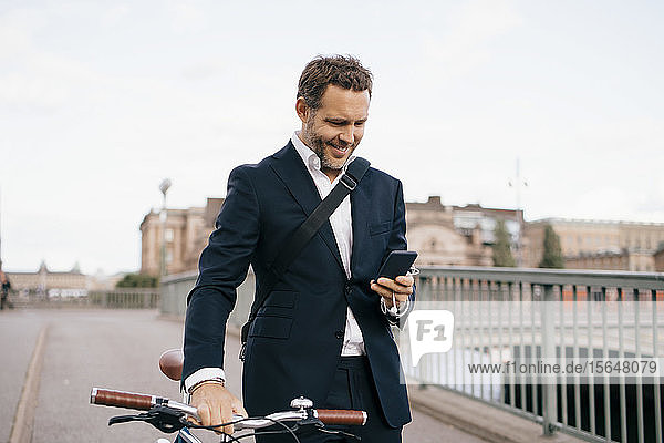 Lächelnder Geschäftsmann mit Mobiltelefon beim Spaziergang mit dem Fahrrad auf einer Brücke in der Stadt