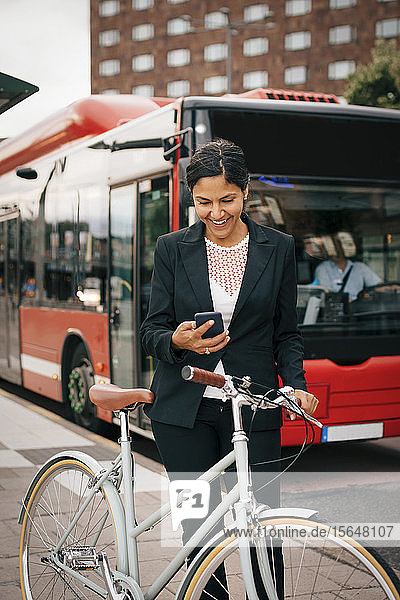 Lächelnde Geschäftsfrau benutzt Smartphone  während sie mit dem Fahrrad gegen den Bus auf der Straße in der Stadt steht