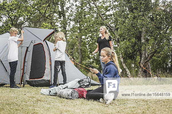 Teenager text messaging on mobile phone while family pitching tent
