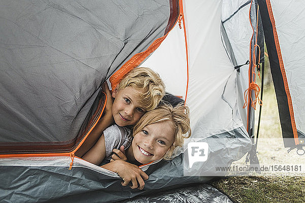 Portrait of smiling siblings peeking through tent at camping site
