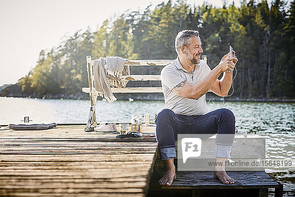 Male wearing headphones photographing through digital tablet while sitting on pier over lake