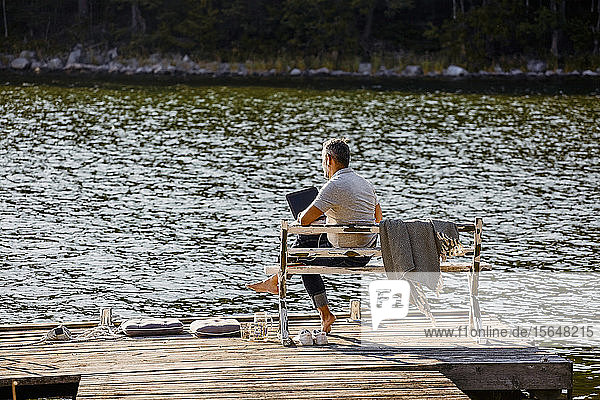 Ein erwachsener Mann sitzt auf einer Bank über einem Holzsteg am See und benutzt einen Laptop