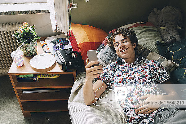 High angle view of smiling teenager listening music through mobile phone on bed at home