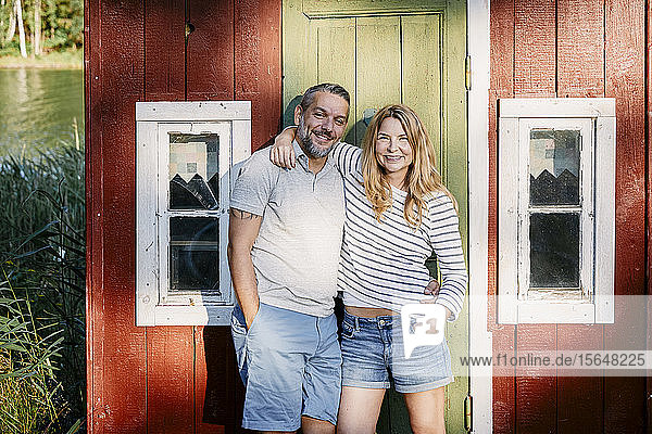 Porträt eines lächelnden Paares  das mit umgebundenen Armen gegen die Tür eines Blockhauses steht