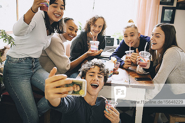 Teenage boy taking selfie with friends on smart phone while drinking smoothie at home