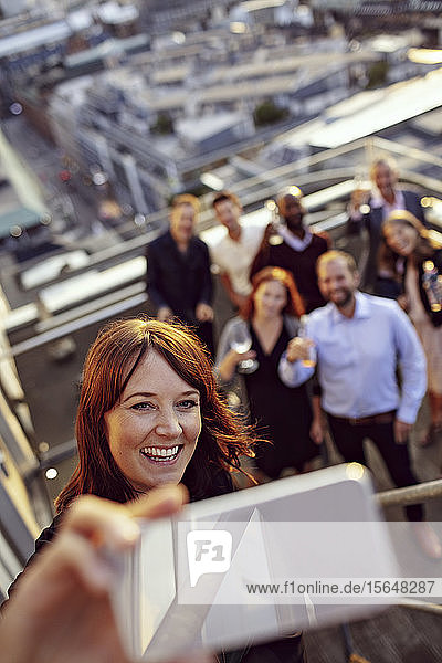 Lächelnde Geschäftsfrau nimmt sich während der Büroparty auf der Terrasse mit Kollegen am Smartphone ein